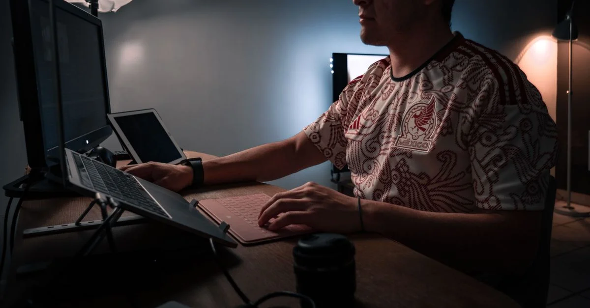 Homme travaillant sur un ordinateur de bureau et un ordinateur portable dans un bureau faiblement éclairé.