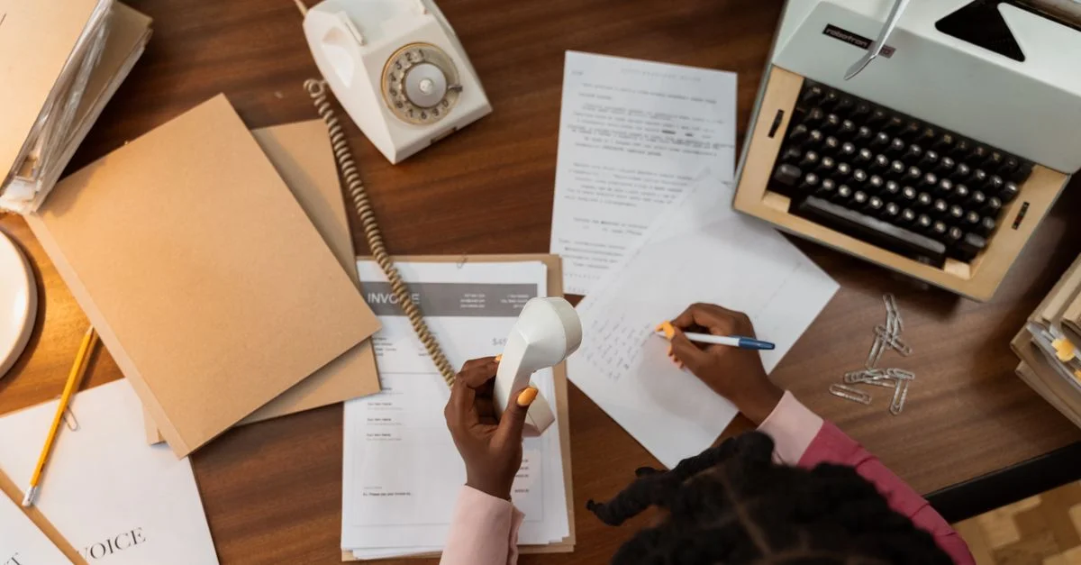 Un bureau de style vintage avec un typewriter, un téléphone et une personne écrivant sur du papier.